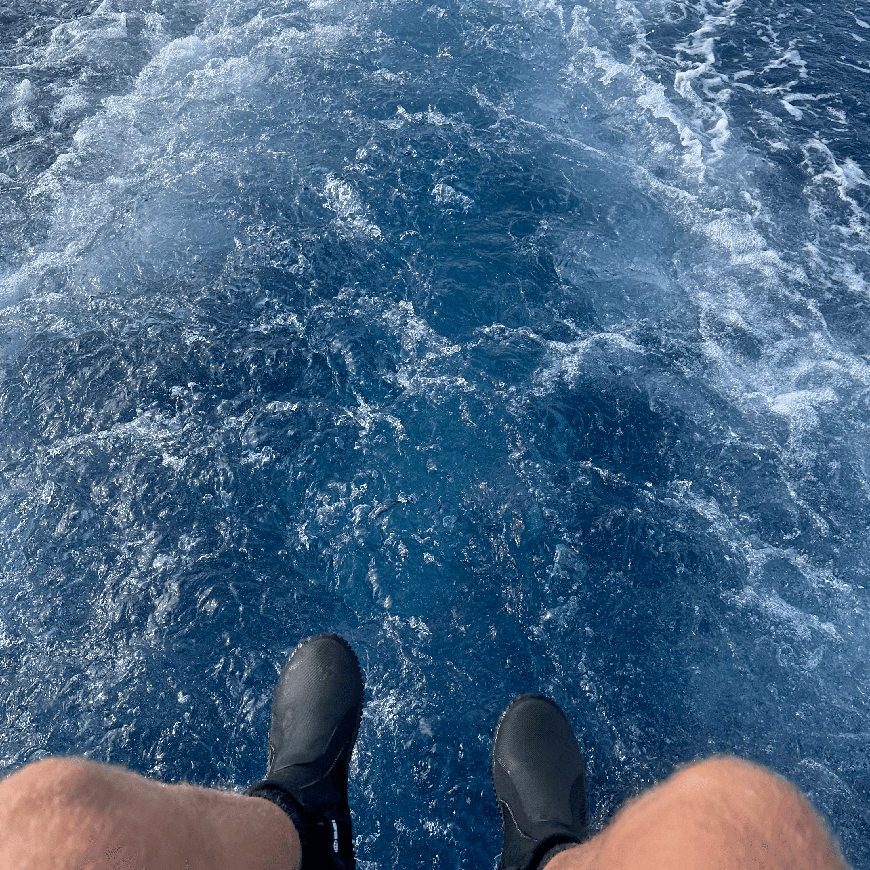 Feet with dive boots dangling off the back of boat over blue ocean water